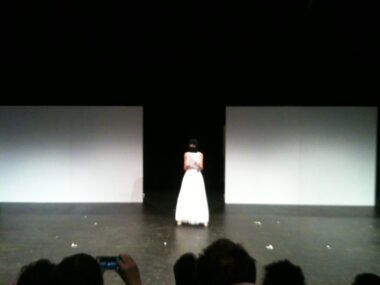 Person center stage in a black ski mask and white ball gown